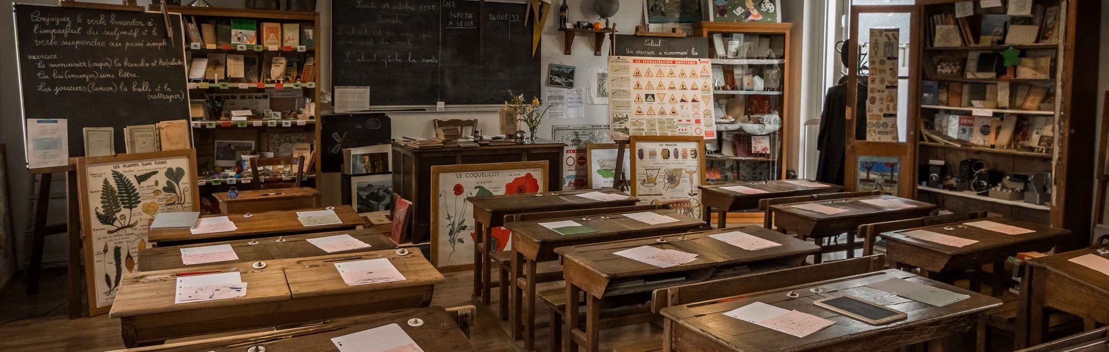 Reconstitution d'une classe des années 50 avec ses tableaux noirs, ses pupitres en bois, ses armoires remplies d'objets scolaires. Les visiteurs accueillis par 1